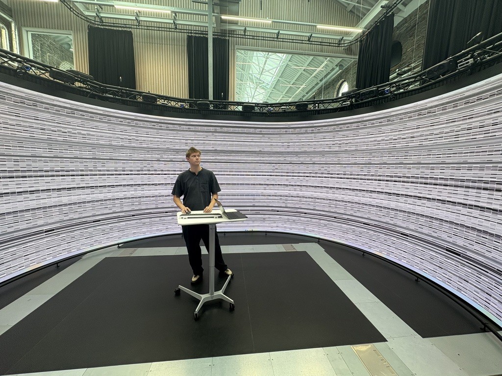 Noah O'Sullivan standing inside the Reality Emulator with an early greyscale iteration of a generative visual on the panoramic screen.
